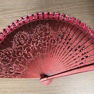 May include: A decorative hand fan with a burgundy and pink color scheme. The fan features an intricate floral design and a pink border. The fan is open, displaying its full shape and design. The fan is made of a material that appears to be paper or fabric.