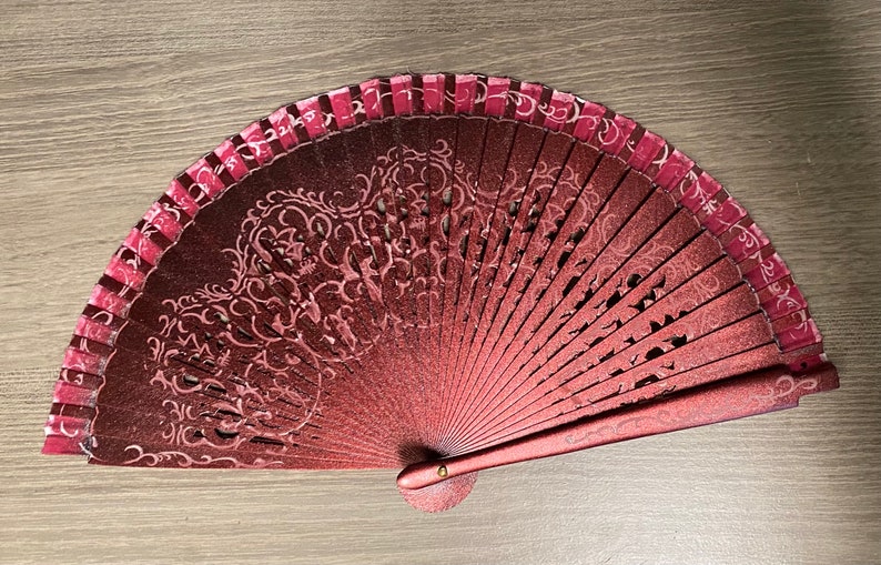 May include: A decorative hand fan with a burgundy and pink color scheme. The fan features an intricate floral design and a pink border. The fan is open, displaying its full shape and design. The fan is made of a material that appears to be paper or fabric.