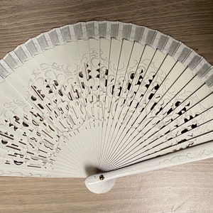 May include: A white folding fan with a decorative design. The fan features a series of vertical panels with cut-out patterns and a decorative border. The handle is white with a small gold-colored rivet.