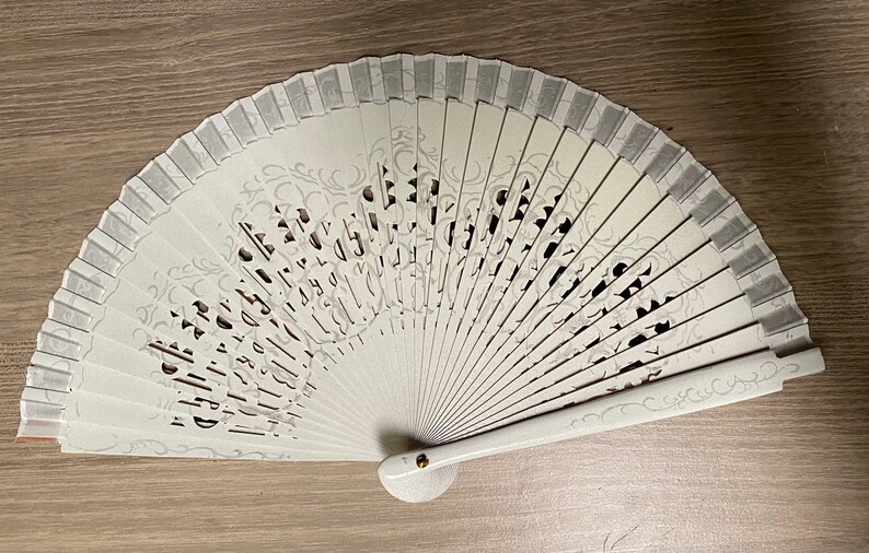 May include: A white folding fan with a decorative design. The fan features a series of vertical panels with cut-out patterns and a decorative border. The handle is white with a small gold-colored rivet.