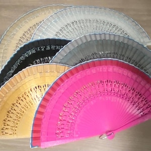 Może przedstawiać: Zestaw sześciu ozdobnych składanych wachlarzy w różnych kolorach, w tym czarny, złoty, szary, różowy i biały. Wachlarze są wykonane z drewna i mają misterne wzory.