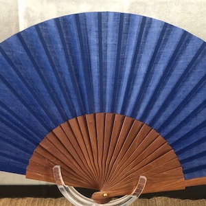 May include: A blue silk folding fan with a wooden frame. The fan is closed and standing upright on a clear stand.