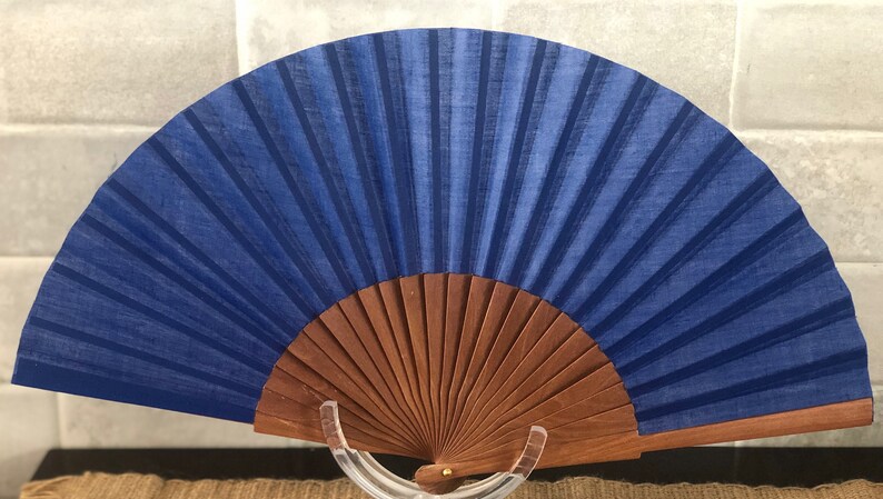 May include: A blue silk folding fan with a wooden frame. The fan is closed and standing upright on a clear stand.