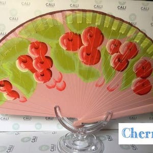 Puede incluir: Un abanico de mano rosa con un dise&ntilde;o de cerezas. El abanico est&aacute; pintado con hojas verdes y cerezas rojas.