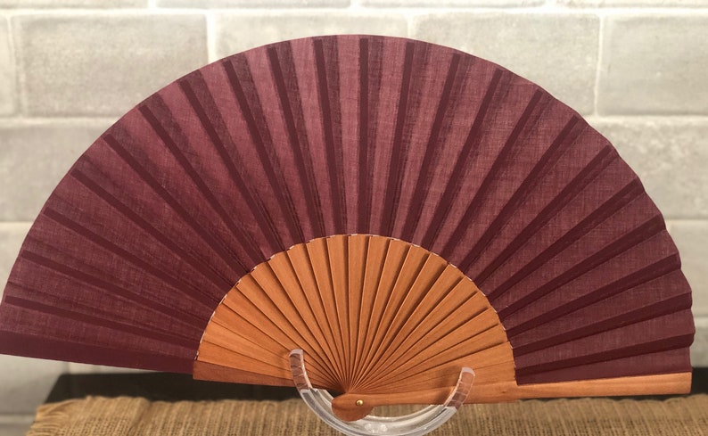 May include: A burgundy hand fan with a wooden frame. The fan is closed and standing on a clear stand.