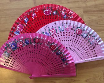 May include: Three hand fans in shades of red and pink, adorned with floral designs. The fans are fanned out, showcasing their painted details. The wooden background provides a neutral backdrop for the vibrant colors.