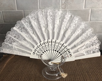 May include: White lace fan with a white wooden handle. The fan is on a clear glass stand.