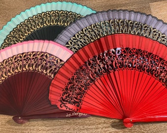 May include: Four decorative folding fans with intricate gold designs on a wooden background. The fans are in various colors, including red, pink, black, and teal.