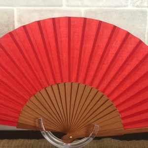 May include: A red hand fan with a wooden frame. The fan is closed and standing on a clear plastic stand.