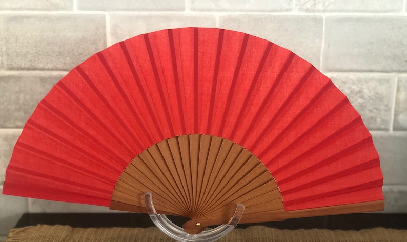 May include: A red hand fan with a wooden frame. The fan is closed and standing on a clear plastic stand.