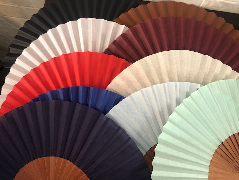 May include: A collection of seven folding fans in various colors, including black, white, red, blue, light blue, and green. The fans are arranged in a semi-circle, with the handles facing outward.