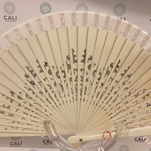 May include: A cream-colored folding hand fan with a decorative, openwork design. The fan features a light beige color with a striped border and a handle. The fan is displayed against a background with the text "CALI ACCESSORIES".