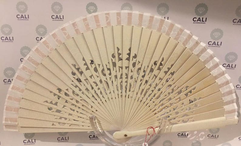 May include: A cream-colored folding hand fan with a decorative, openwork design. The fan features a light beige color with a striped border and a handle. The fan is displayed against a background with the text "CALI ACCESSORIES".