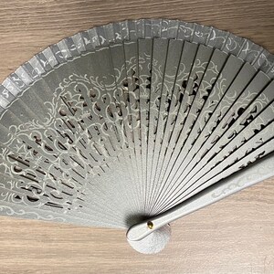 May include: A gray hand fan with a decorative, openwork design. The fan features a silver-colored, ornate pattern and a small, round base. The fan is made of wood and has a metallic finish. The fan is open and fanned out.