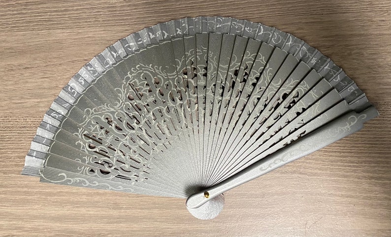 May include: A gray hand fan with a decorative, openwork design. The fan features a silver-colored, ornate pattern and a small, round base. The fan is made of wood and has a metallic finish. The fan is open and fanned out.