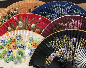 May include: A collection of hand-painted folding fans in various colors. The fans feature floral designs in shades of yellow, white, red, blue, and purple. The fans are made of wood and fabric, and are arranged in a semi-circle.