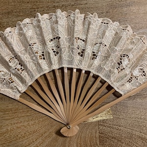 May include: A white lace fan with a wooden handle and a gold tassel. The fan is closed and has a delicate floral pattern.