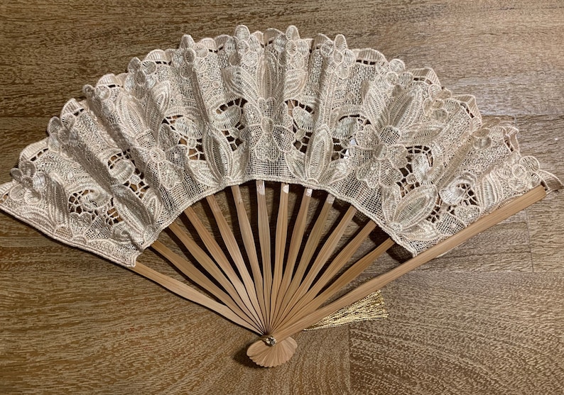 May include: A white lace fan with a wooden handle and a gold tassel. The fan is closed and has a delicate floral pattern.
