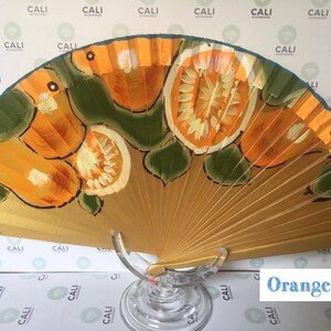 Puede incluir: Un abanico plegable de madera pintado a mano con fondo dorado y un dise&ntilde;o de rodajas de naranja y hojas verdes. El abanico est&aacute; sobre un soporte de vidrio transparente. El texto "Orange 2" es visible en la esquina inferior derecha de la imagen.