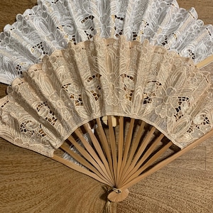 May include: A set of two decorative folding fans with a wooden frame and lace fabric. The fans are in white and beige with a floral pattern. The fan has a gold tassel.