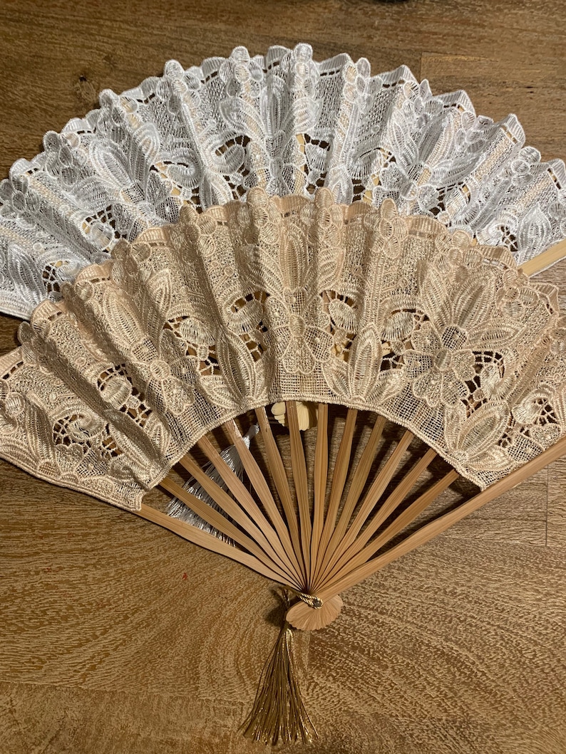 May include: A set of two decorative folding fans with a wooden frame and lace fabric. The fans are in white and beige with a floral pattern. The fan has a gold tassel.