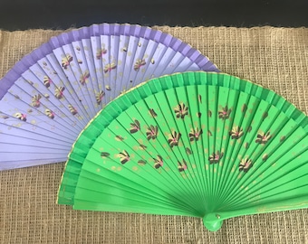 May include: Two hand fans, one is light purple with gold and pink floral designs, the other is bright green with gold and pink floral designs.