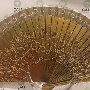 May include: A decorative hand fan with a gold and brown color scheme. The fan features an intricate, openwork design with gold accents and a silver border. The fan is displayed on a clear stand.