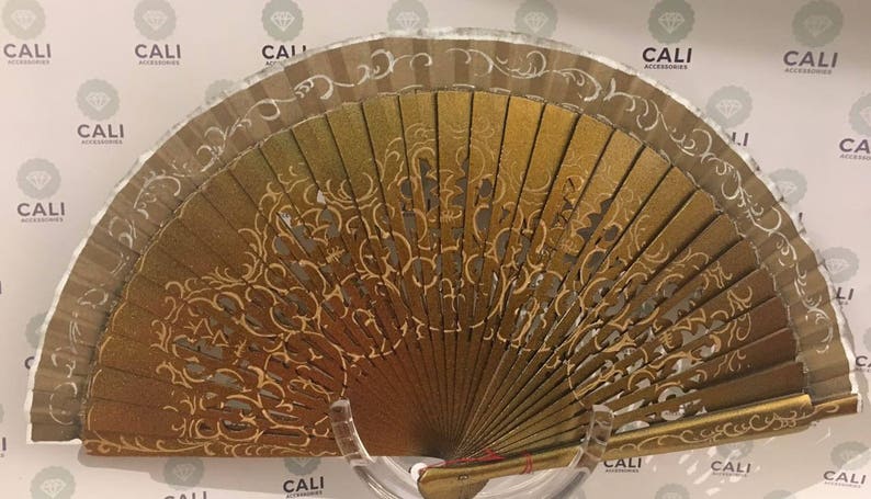 May include: A decorative hand fan with a gold and brown color scheme. The fan features an intricate, openwork design with gold accents and a silver border. The fan is displayed on a clear stand.