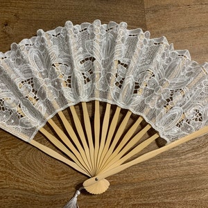 May include: A white lace folding fan with a wooden handle and a tassel. The fan is closed and the lace design is intricate.
