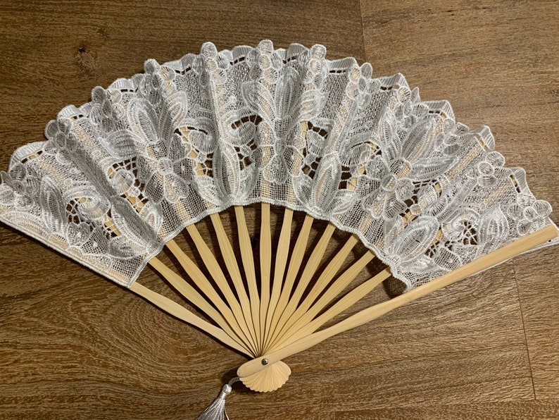 May include: A white lace folding fan with a wooden handle and a tassel. The fan is closed and the lace design is intricate.