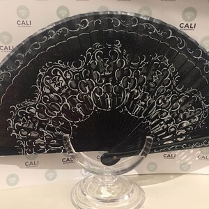 May include: A black hand fan with a white, ornate design. The fan is open, displaying a detailed pattern of swirls and floral motifs. The fan is on a clear stand, and the background has the word "CALI" repeated.