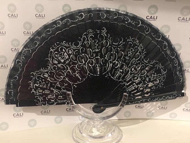 May include: A black hand fan with a white, ornate design. The fan is open, displaying a detailed pattern of swirls and floral motifs. The fan is on a clear stand, and the background has the word "CALI" repeated.