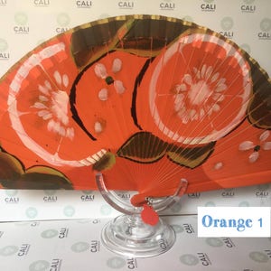 Puede incluir: Un abanico de mano rojo y dorado con un dise&ntilde;o pintado de dos rodajas de naranja. El abanico est&aacute; en un soporte transparente. El texto "Orange 1" es visible en la esquina inferior derecha de la imagen.