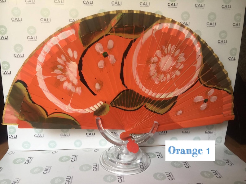 Puede incluir: Un abanico de mano rojo y dorado con un dise&ntilde;o pintado de dos rodajas de naranja. El abanico est&aacute; en un soporte transparente. El texto "Orange 1" es visible en la esquina inferior derecha de la imagen.