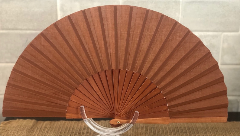 May include: A brown wooden hand fan with a decorative design. The fan is closed and is on a clear stand.