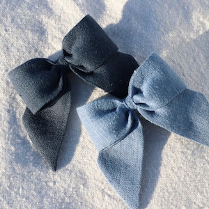 May include: Three handmade fabric bows in shades of blue. The bows are made of textured linen fabric. One bow is a dark navy blue, another is a lighter denim blue, and the third is a medium blue. The bows are arranged on a light-colored, textured surface.