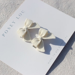May include: Two off-white linen hair bows on a white card. The bows are tied in a classic bow shape and attached to silver-colored metal clips. The card has the text "POSEY LOU" and "made in gig harbor, wa".