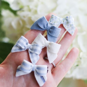 May include: Five small fabric hair bows in various patterns and colors, including blue, white, and floral prints. The bows are arranged on a light background, showcasing their delicate details and textures. Each bow is approximately 2 inches wide.