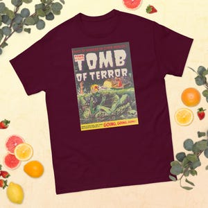 以下が含まれることがあります： マロン色のTシャツに、ビンテージコミックの表紙のデザインがプリントされています。表紙には、水から現れたモンスターと、「Tomb of Terror」と「Going, Going, Gone!」の文字が書かれています。