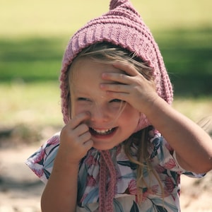 May include: A dusty rose knitted bonnet with a pointed top and ribbed texture, featuring long braided ties that hang down. The bonnet is paired with a short-sleeved shirt that has a white base and a repeating pattern of pink and blue floral elements.