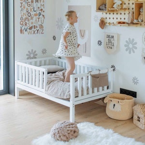 Puede incluir: Una cama de niño pequeña de color blanco con un marco de madera blanco y un colchón blanco. La cama tiene una barandilla de madera blanca en un lado. Una niña pequeña está saltando en la cama. La pared detrás de la cama está decorada con flores blancas y el texto "Dream Big Little One".