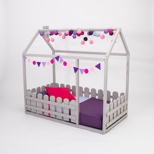 Peut inclure: Un lit en bois gris en forme de maison avec un matelas à motifs violets et roses et un oreiller rose. Le cadre de lit est doté d'une clôture en bois blanc autour du matelas et d'une guirlande de lumières violettes et roses suspendues au sommet.