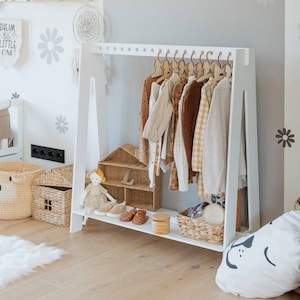 May include: White wooden clothes rack with a white shelf and a white background. The rack is filled with children's clothing on wooden hangers. There are two wicker baskets on the shelf, one with a doll and the other with toys. The rack is in a room with a white rug and a white pillow with a black dog design.