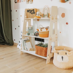 May include: A light-colored wooden bookshelf with three shelves, holding books, toys, and a decorative basket. The bookshelf has a ladder-like design. A large, knitted basket with a bear face sits on the floor.