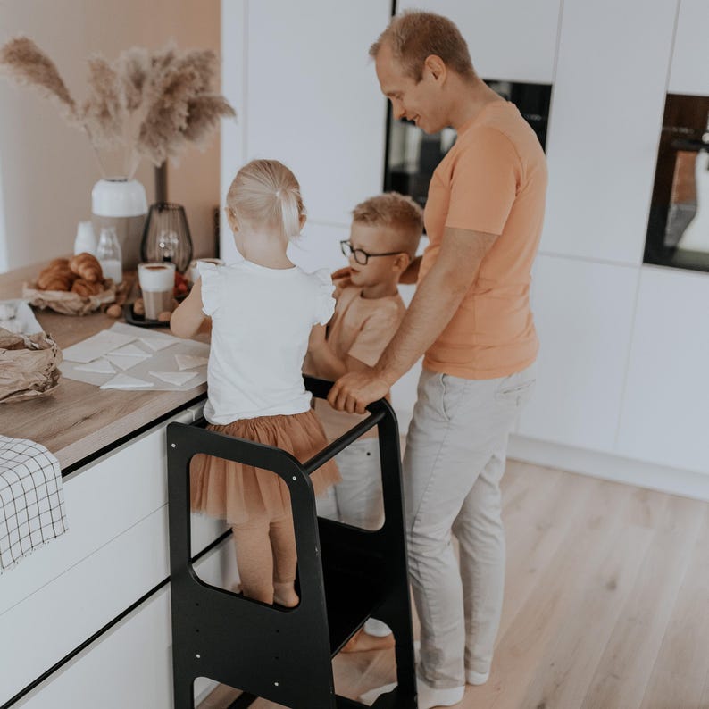 Father assisting two kids standing on a matte black kitchen step stool, giving grandmothers a functional Name Day surprise idea.