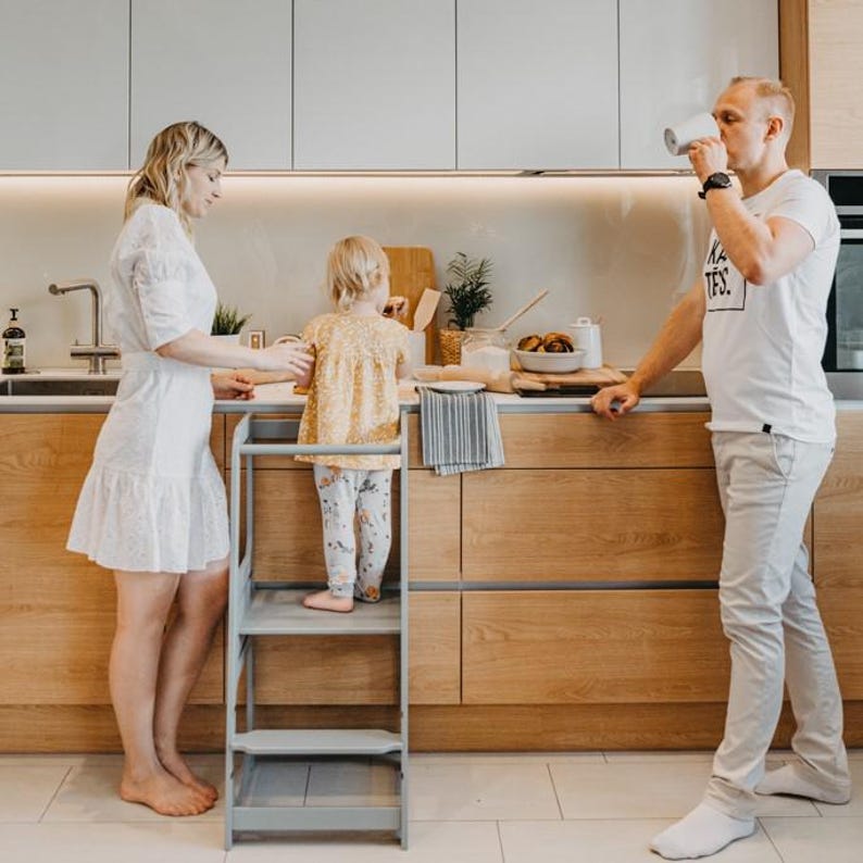 Grey montessori toddler kitchen step stool supporting a young boy at the island while parents enjoy morning coffee.