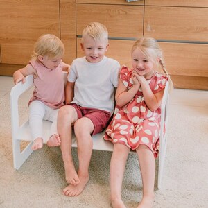 Three smiling young children sitting side-by-side on a white wooden double bench.
