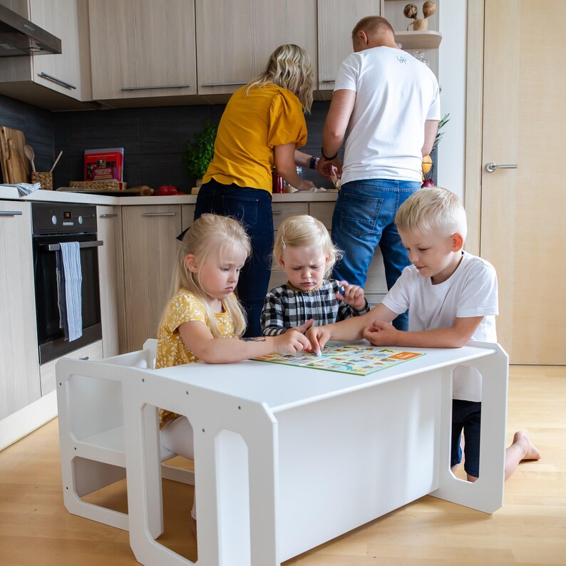 Children playing with a board game at a white toddler play desk with family in the background.