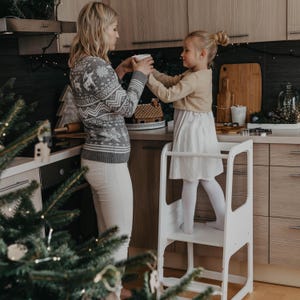 Mom giving a warm drink to her toddler standing on a white step stool in a holiday decorated kitchen, ideal for Christmas morning.