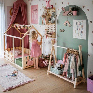 Toddler girl hanging pastel dresses on a wooden rack near a timber house bed, creating an organized capsule wardrobe layout for young girls.